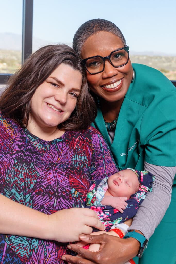 Two women smiling while holding a newborn baby, with one woman wearing medical scrubs in a hospital setting.