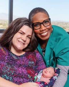 Two women smiling while holding a newborn baby, with one woman wearing medical scrubs in a hospital setting.