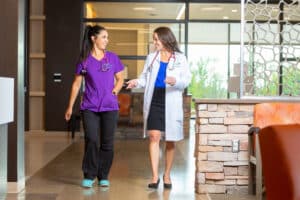 Two women smiling while holding a newborn baby, with one woman wearing medical scrubs in a hospital setting.